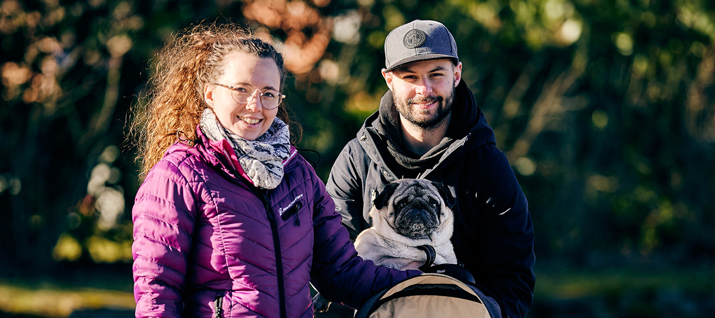 Ett par är ute med barnvagnen i solen och bär en hund.